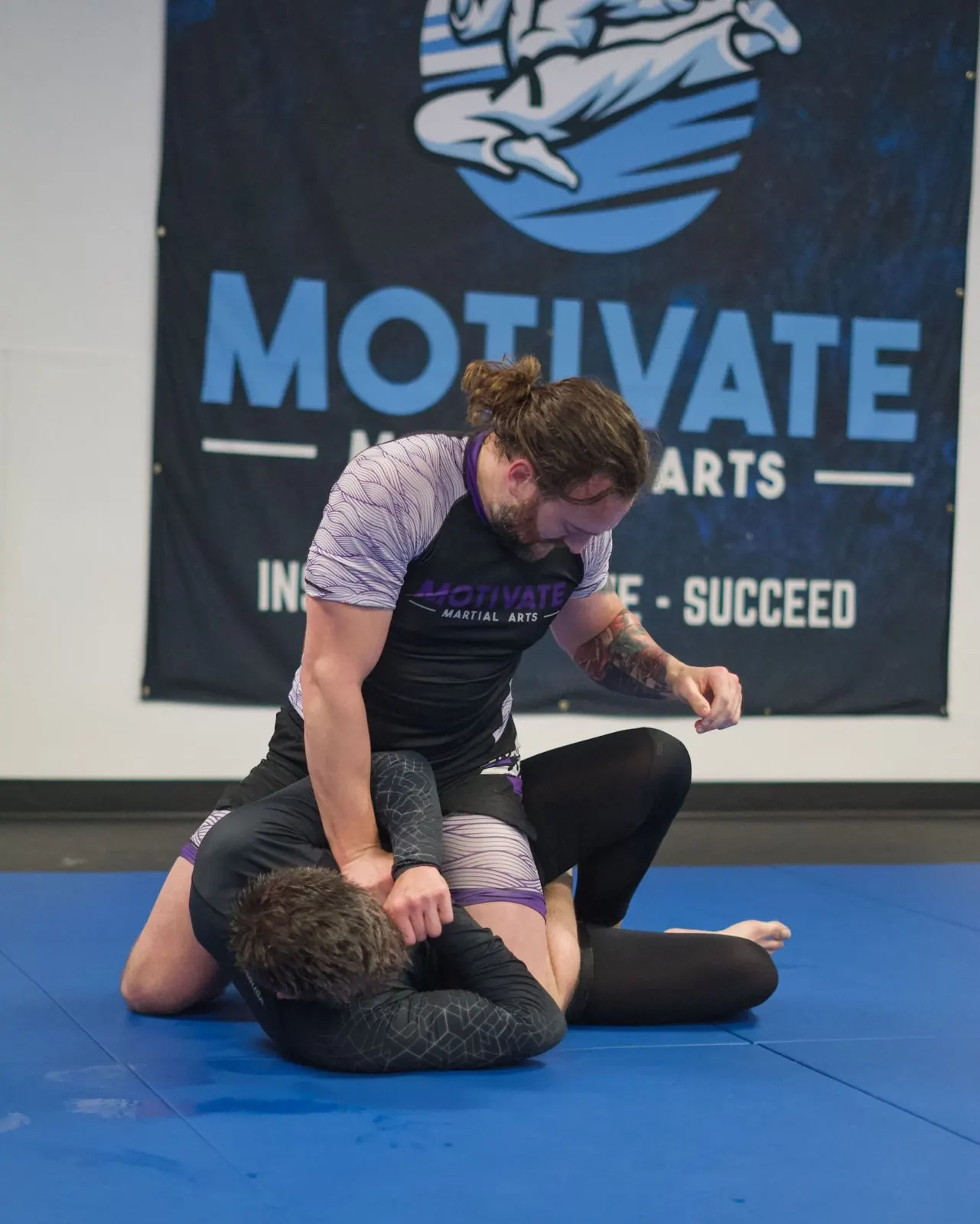Two adults training No-Gi Brazilian Jiu-Jitsu in rashguards on the blue mats