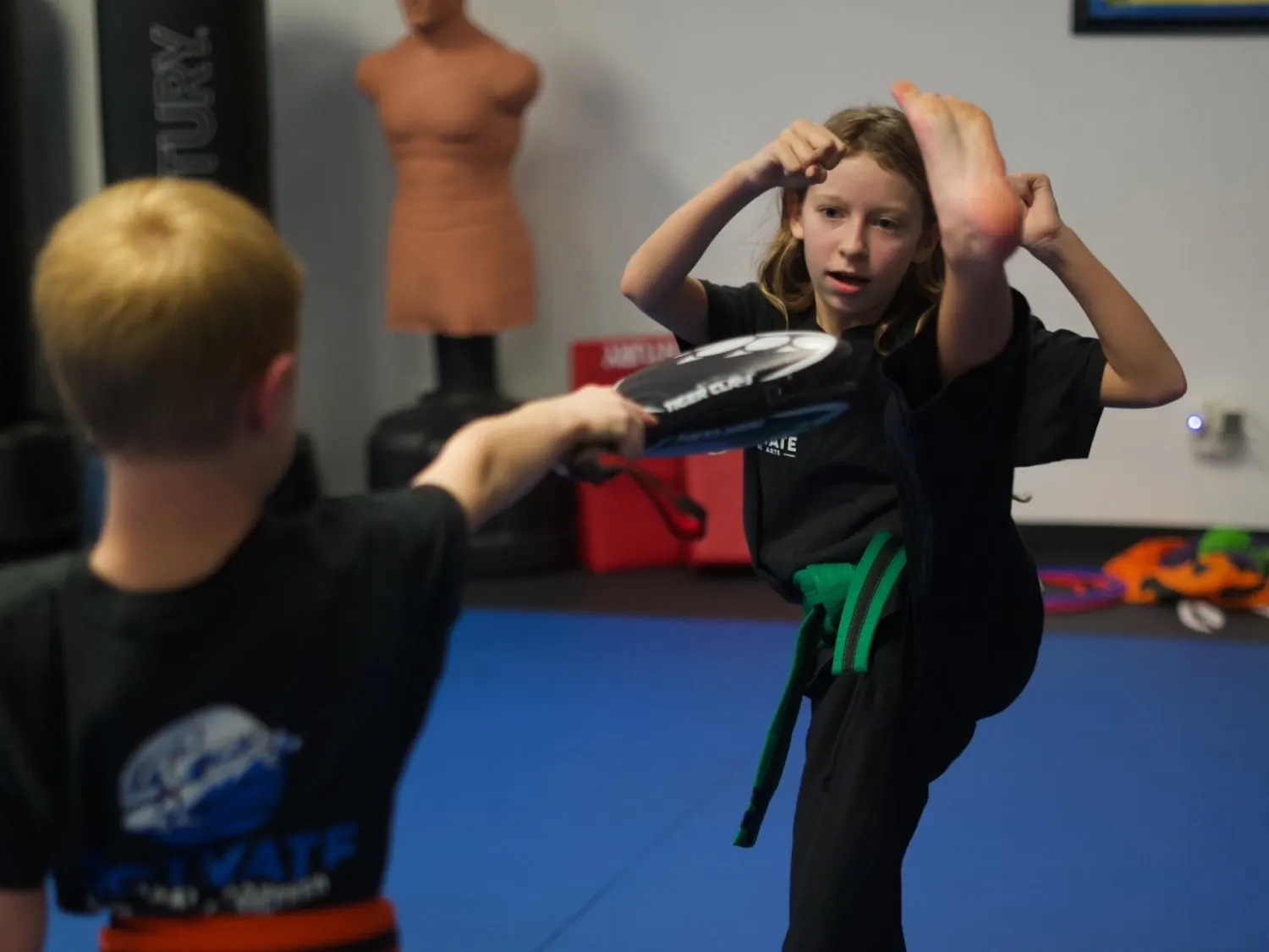 A green-belt Junior TaeKwonDo student landing a high kick on a target paddle