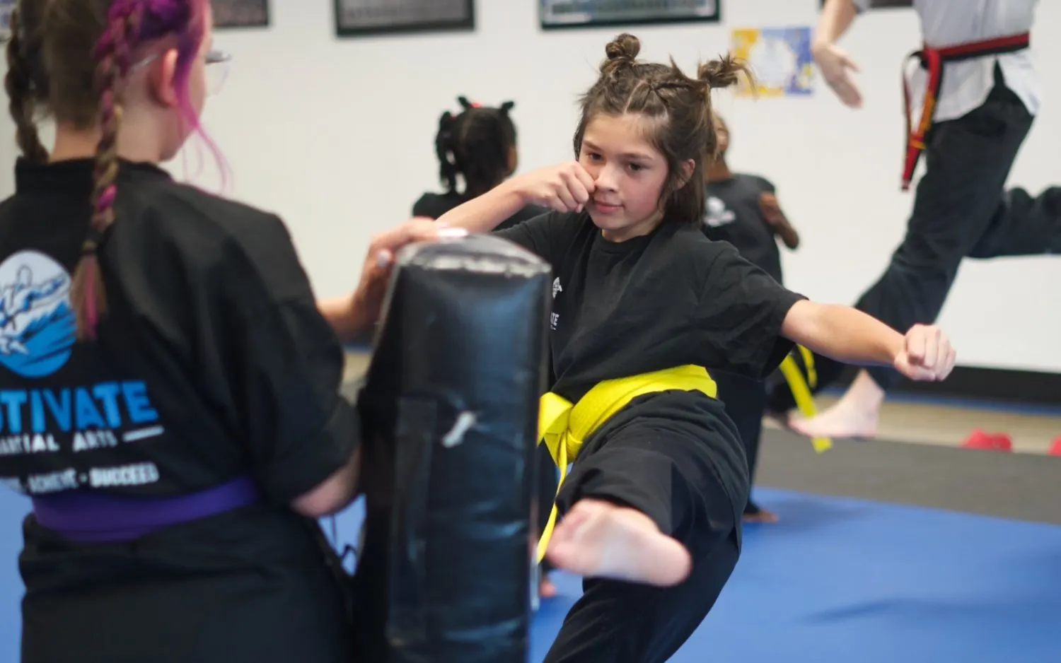 A yellow-belt Junior TaeKwonDo student kicking a kick shield held by her partner on the blue mats