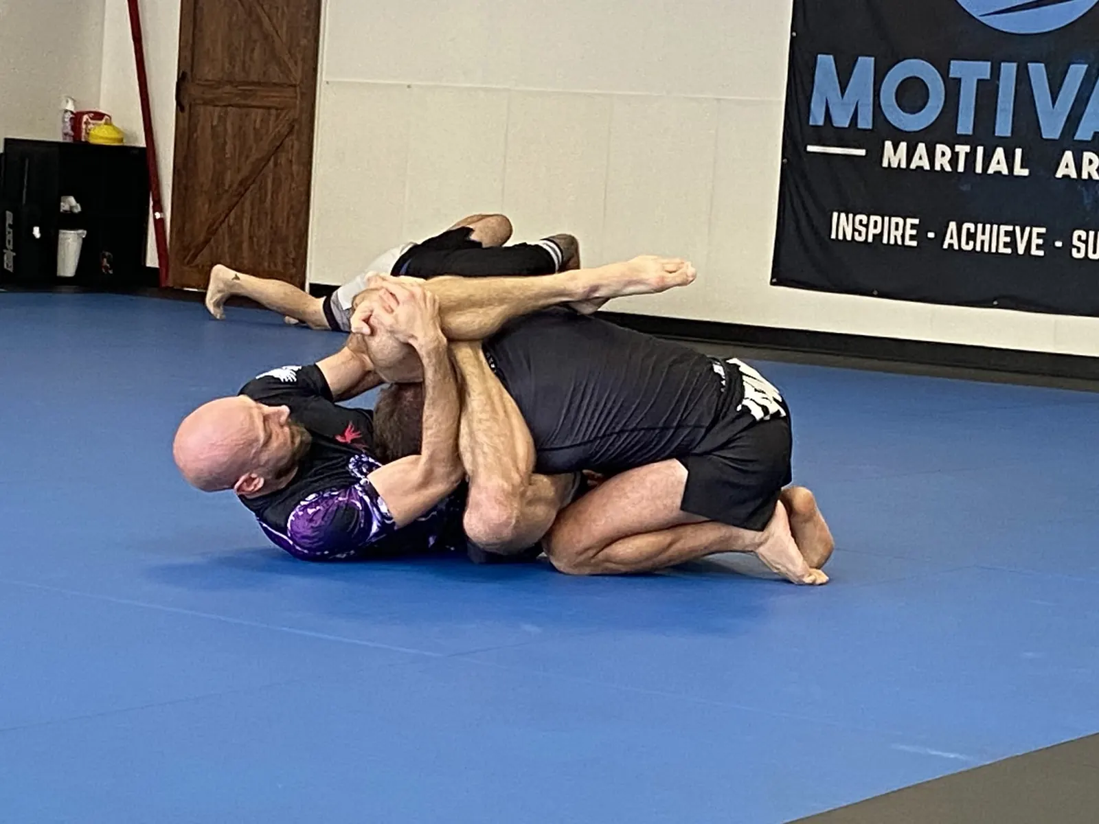 Two adults grappling No-Gi in rashguards on the blue mats in front of the Motivate banner