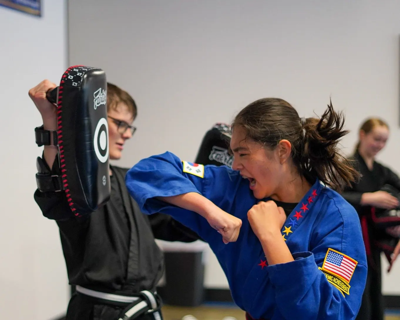 A teen TaeKwonDo student in a blue uniform striking a kick pad held by a partner